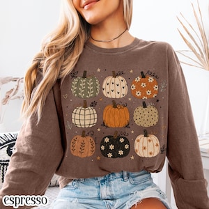 May include: A brown long-sleeve sweatshirt featuring a graphic print of nine pumpkins in various patterns and colors, including floral, heart, plaid, and polka dot designs, accented with small stars and the word espresso in the corner.