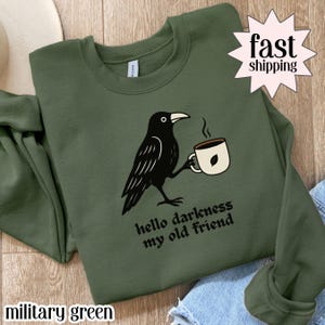May include: Military green sweatshirt featuring a black crow holding a coffee cup with a leaf design. The text "hello darkness my old friend" is printed below the crow. The sweatshirt has a crew neck and long sleeves.