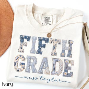 May include: Ivory-colored t-shirt with the words "FIFTH GRADE" in blue patchwork letters. The shirt also features the name "miss taylor" in cursive. A gold necklace with a pendant is draped across the shirt.