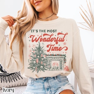 May include: Ivory-colored long-sleeve shirt with a Christmas-themed graphic. The graphic features a decorated Christmas tree, a fireplace, and the text "It's the Most Wonderful Time" in red and green.