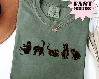 May include: An olive green t-shirt with a crew neck and short sleeves, featuring five black cat illustrations. A silver necklace with a small pendant is visible. The tag reads "Comfort Colors".