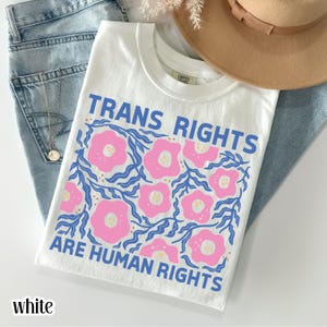 May include: White t-shirt with the text "TRANS RIGHTS ARE HUMAN RIGHTS" in blue. The shirt features a floral design with pink flowers and blue leaves. The shirt is folded on top of blue jeans, with a gold necklace and a brown hat in the background.
