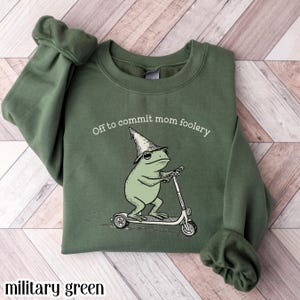 May include: Military green sweatshirt with the text "Off to commit mom foolery." The graphic features a light green frog wearing a wizard hat and riding a scooter. The sweatshirt is a solid colour.
