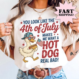 Puede incluir: Camiseta color crema con un gráfico de un pato con gafas de sol y un sombrero, sosteniendo un perrito caliente y una bandera estadounidense. El texto dice: "You look like the 4th of July makes me want a hot dog real bad!"