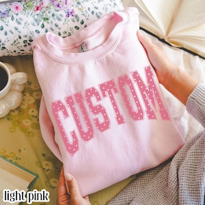 Puede incluir: Una sudadera de cuello redondo en color rosa claro con la palabra CUSTOM impresa en letras grandes, negritas y estilo bloque. Las letras son rosas con un patrón de lunares blancos. La prenda tiene cuello acanalado y mangas largas.