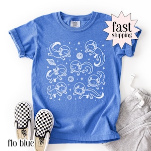 May include: A blue t-shirt featuring a white graphic of axolotls in space suits, planets, and a spaceship. The shirt has a crew neck and short sleeves. The words "fast shipping" are in a starburst graphic. A pair of black and white checkered slip-on shoes are at the bottom.