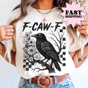 May include: Cream-colored t-shirt featuring a black crow perched on a branch, surrounded by flowers and a checkered pattern. The text "F-CAW-F" is above the crow. The design is a vintage-style illustration.