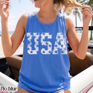 Puede incluir: Una camiseta sin mangas azul con la palabra "USA" impresa en blanco, con un patrón de estrellas dentro de las letras. La camiseta sin mangas es de color azul liso y tiene cuello redondo. El texto "flo.blue" está en la parte inferior izquierda.