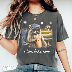 Può includere: T-shirt grigio scuro con un procione con occhiali da sole e un berretto da baseball, che tiene in mano una bevanda e seduto su una sedia blu. La grafica include il testo "i live here now" e uno sfondo di campo da baseball.