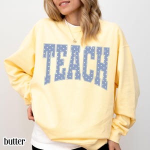 May include: A pale yellow long-sleeve sweatshirt featuring the word TEACH in large, block-style letters. The letters are light blue with a white polka dot pattern. The garment has a crew neckline and a relaxed fit, providing a simple design.