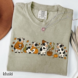Puede incluir: Una camiseta de manga corta color caqui con un estampado gráfico de cuatro vacas de dibujos animados con atuendos otoñales, incluyendo suéteres, bufandas y gorros, rodeadas de calabazas, hojas y cestas. La camiseta tiene una etiqueta Comfort Colors.