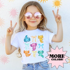 Könnte beinhalten: Ein weißes T-Shirt mit einem bunten Design aus Axolotln mit Schleifen. Die Axolotl sind in verschiedenen Farben, darunter Blau, Rosa, Gelb und Orange. Der Text "MEHR FARBEN!" befindet sich in einem rosa Sternenausbruch.