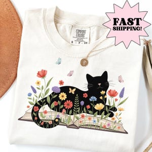 Ivory Comfort Colors graphic t-shirt featuring a black cat resting on an open book surrounded by colorful wildflowers and butterflies, cozy bookish cat shirt for readers and cat lovers, soft unisex floral cat tee perfect for librarians, teachers, bookworms, gifts, and everyday casual wear