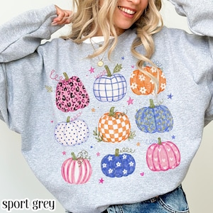 May include: A light gray crewneck sweatshirt featuring a graphic print of nine pumpkins in various patterns and colors, including leopard print, plaid, checkered, floral, and striped designs, accented with small stars and vines.