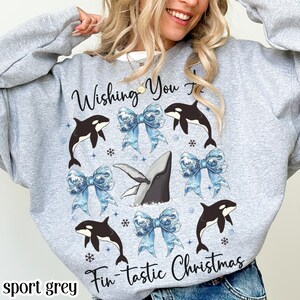 May include: Sport grey sweatshirt with a festive design. The design features black and white orcas, blue bows, snowflakes, and a whale. Text reads "Wishing You a Fin-tastic Christmas".