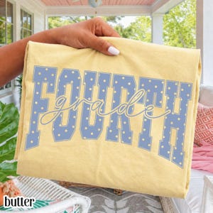 May include: A pale yellow t-shirt featuring the text "FOURTH grade" in a large, blocky, light blue font with white polka dots. The word "FOURTH" is printed in a bold, collegiate-style typeface, while "grade" is written in a cursive script.