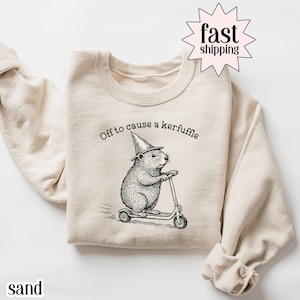 May include: Sand-colored sweatshirt with a whimsical illustration of a rodent wearing a wizard hat and riding a scooter. The text "Off to cause a kerfuffle" is printed above the rodent. The word "sand" is printed at the bottom left.