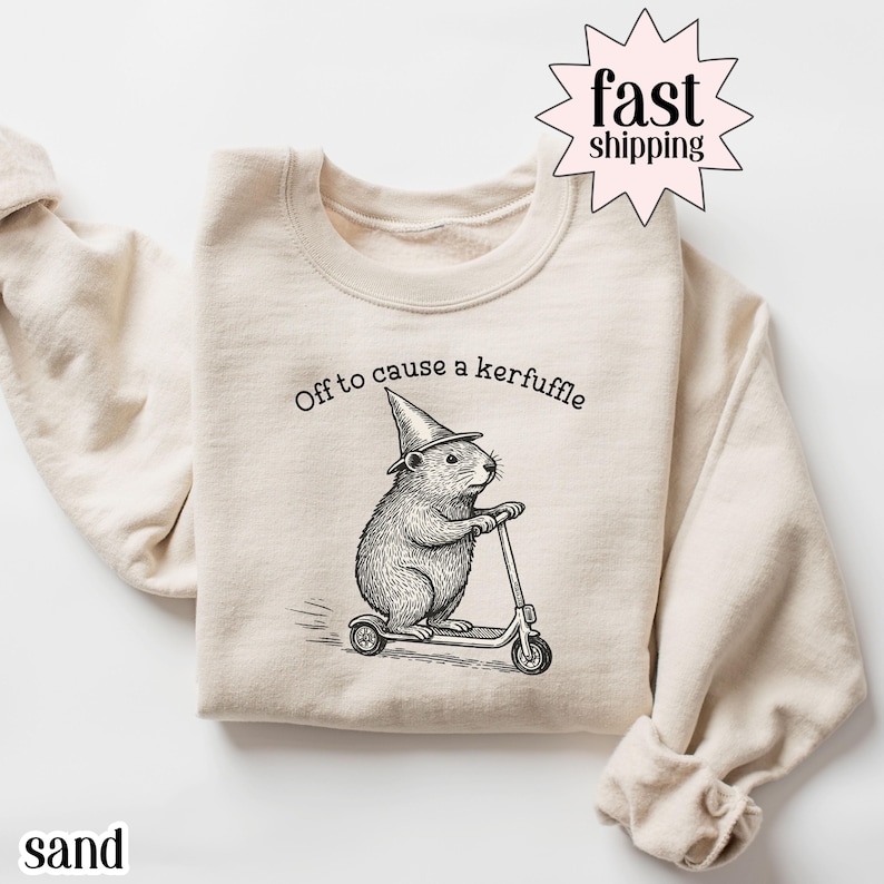 May include: Sand-colored sweatshirt with a whimsical illustration of a rodent wearing a wizard hat and riding a scooter. The text "Off to cause a kerfuffle" is printed above the rodent. The word "sand" is printed at the bottom left.