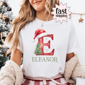 Puede incluir: Camiseta blanca con un diseño festivo. La letra "E" roja está adornada con copos de nieve y un gorro de Papá Noel. Un pequeño árbol de Navidad está al lado de la letra. El nombre "Eleanor" está impreso en verde.