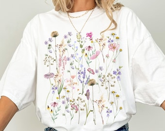 May include: White t-shirt featuring a colorful floral design. The graphic includes various wildflowers in shades of pink, purple, yellow, and green. The word "white" is printed at the bottom left corner of the shirt.