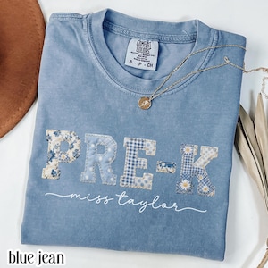 Może przedstawiać: Niebieska koszulka Comfort Colors z napisem "PRE-K miss taylor" w naszytych literach. Litery są w różnych niebieskich kwiatowych i kratowych wzorach. Złoty naszyjnik z wisiorkiem spoczywa na koszulce.