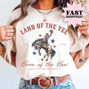 Cream graphic shirt featuring a western-style patriotic illustration of a cowboy riding a bucking horse with red and blue stars and text reading “Land of the Free” and “Home of the Brave.” Patriotic western graphic tee designed as Americana apparel, rodeo-inspired shirt, and USA pride statement tee