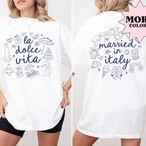 以下が含まれることがあります： 白いTシャツに、青いグラフィックデザインが施されています。デザインには「la dolce vita」というテキストと、イタリアをテーマにした様々なイラストが描かれています。Tシャツの裏面には、青いグラフィックデザインが施されており、「イタリアで結婚」というテキストと、イタリアをテーマにした様々なイラストが描かれています。