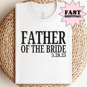 Może przedstawiać: Biała koszulka z napisem "FATHER OF THE BRIDE" pogrubioną czarną czcionką, wraz z datą "5.28.25". Koszulka jest prezentowana na tkanej powierzchni. Różowa grafika w kształcie gwiazdy z napisem "FAST SHIPPING!" znajduje się w prawym górnym rogu.