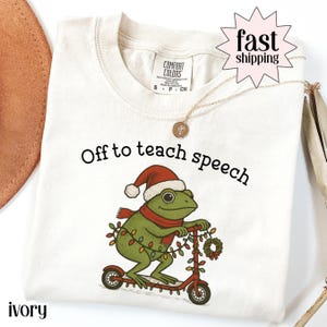 Op de afbeelding: Ivoor t-shirt met de tekst "Off to teach speech". Het ontwerp toont een cartoon kikker met een kerstmuts en sjaal, rijdend op een rode scooter versierd met kerstverlichting en een krans. Een gouden ketting is ook zichtbaar.