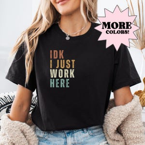 May include: Black t-shirt with a vintage-style graphic that reads "IDK I JUST WORK HERE".