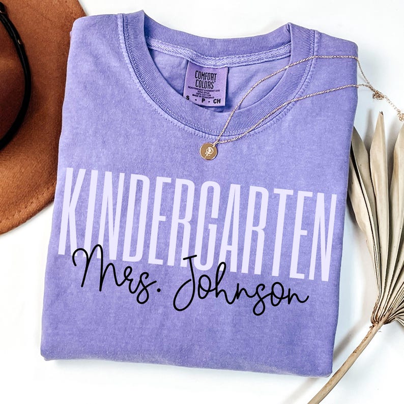May include: A lavender Comfort Colors t-shirt with the words "KINDERGARTEN Mrs. Johnson" printed in white and black. A gold necklace with a pendant rests on the shirt. A brown hat and dried plant are in the background.