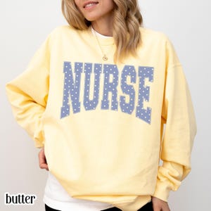 May include: A pale yellow long-sleeve sweatshirt featuring the word NURSE printed in a large, arched, collegiate-style font. The letters are light blue with a white polka dot pattern throughout, creating a distinct contrast against the yellow fabric.