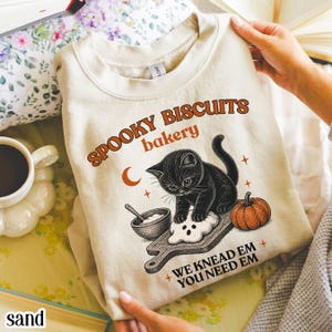 Puede incluir: Una sudadera de cuello redondo color arena con un estampado gráfico de un gatito negro amasando masa sobre una tabla de madera. El texto dice SPOOKY BISCUITS bakery y WE KNEAD EM YOU NEED EM, junto a una pequeña calabaza naranja y una luna.