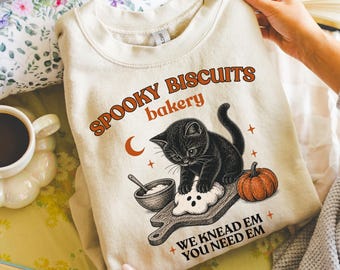 May include: A sand-colored crewneck sweatshirt featuring a graphic print of a black kitten kneading dough on a wooden board. The text reads SPOOKY BISCUITS bakery and WE KNEAD EM YOU NEED EM, accompanied by a small orange pumpkin and crescent moon.