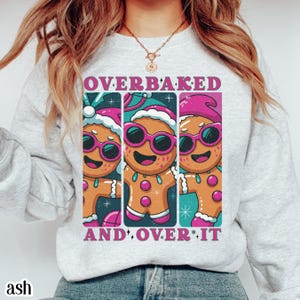 May include: Ash-colored sweatshirt with a graphic of three gingerbread men wearing sunglasses and Santa hats. The text "OVERBAKED AND OVER IT" is printed above and below the gingerbread men. The sweatshirt is a crew neck style.