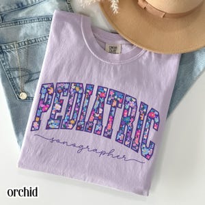 Puede incluir: Una camiseta lila con la palabra "PEDIATRIC" en un diseño floral y la palabra "sonographer" en cursiva. La camiseta está doblada sobre unos vaqueros azules, con un collar dorado y un sombrero beige en el fondo.
