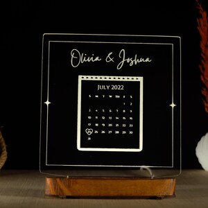 May include: A white acrylic calendar with a black base, featuring the names "Olivia & Joshua" and a calendar for July 2022.