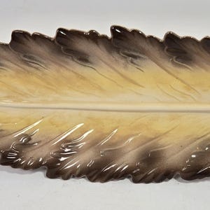 May include: A ceramic serving tray shaped like an elongated leaf with a central vein. The piece features a gradient color scheme, transitioning from a light cream center to dark brown, scalloped edges with a glossy, smooth finish.