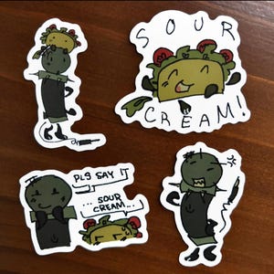 May include: Four quirky stickers with cartoon tacos and characters. One sticker says "SOUR CREAM!" and another says "PLS SAY IT ... SOUR CREAM...". The tacos have smiling faces and are decorated with red and green toppings.