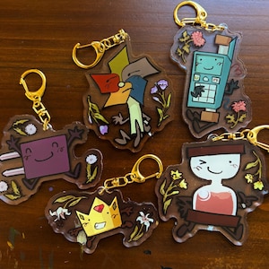 以下が含まれることがあります： ゴールドカラーの金具が付いた、6つのアクリルキーホルダーのコレクション。各キーホルダーは、花のアクセントが付いたユニークな漫画のキャラクターデザインが特徴です。キャラクターは、紫、黄色、青など、さまざまな色です。
