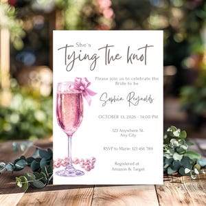 Pode incluir: Um cartão branco com uma taça de champanhe rosa e um laço rosa. O texto no cartão diz "She's Tying the Knot" e inclui o nome da noiva, Sophia Reynolds, a data e hora do evento, o local, as informações de RSVP e onde o casal está registrado.