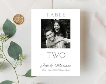 Może przedstawiać: Czarno-biała karta numeru stołu weselnego z tekstem "TABLE TWO" i imionami "Jake & Madison" z datą "25th July 2022, Miami Beach". Czarno-białe zdjęcie pary znajduje się w środku karty.