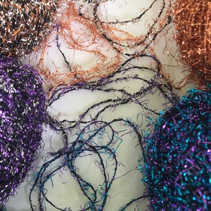 May include: Close-up of various metallic yarn balls and strands in shades of purple, copper, and black. The yarn has a shiny, textured appearance, with some strands intertwined and others forming loose balls. The background is a soft, white surface.