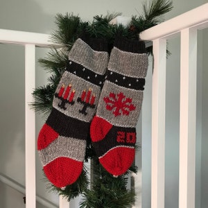 May include: Two gray and black knitted Christmas stockings with red accents. One stocking features a snowflake design and the other features a candle design. The stockings are hanging on a white railing.