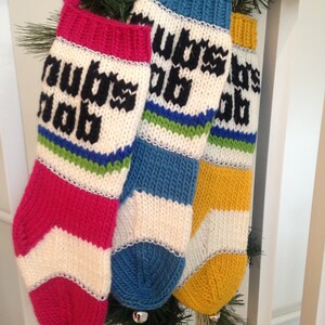 May include: Three knitted Christmas stockings with white, red, blue, and yellow stripes. The stockings are decorated with silver trim and have the words "Now is the" on the red stocking, "Wassail" on the blue stocking, and "Time to" on the yellow stocking.