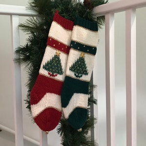 May include: Two knit Christmas stockings, one with red accents and one with dark green accents, both featuring a cream-colored center panel with a green Christmas tree motif, gold star embellishments, and a small metallic bell at the toe.
