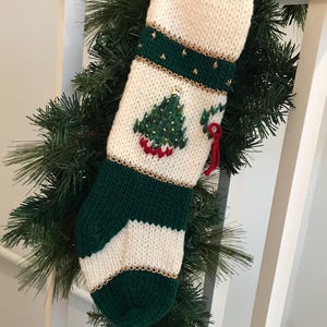 以下が含まれることがあります： 金色の縁取りと小さな刺繍されたクリスマスツリーが付いた緑と白のニット製のクリスマスストッキング。ストッキングの底にはベルが付いています。