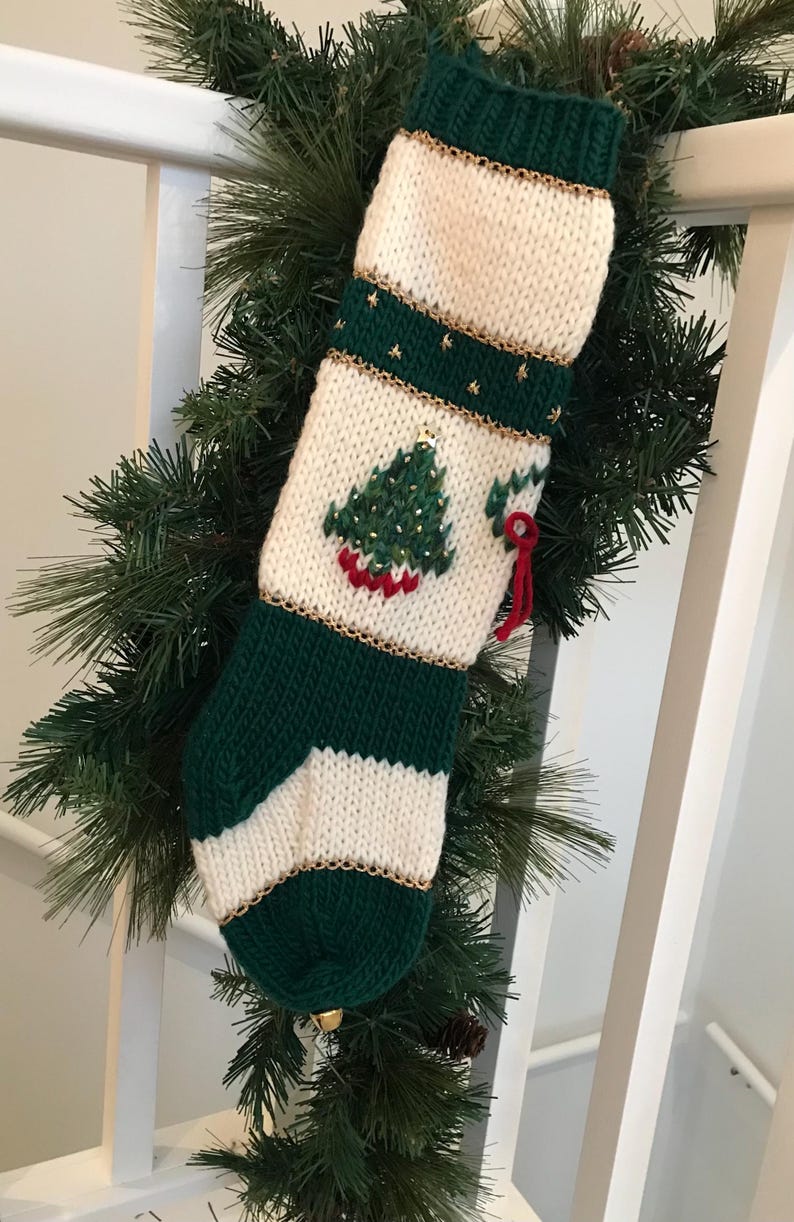 以下が含まれることがあります： 金色の縁取りと小さな刺繍されたクリスマスツリーが付いた緑と白のニット製のクリスマスストッキング。ストッキングの底にはベルが付いています。
