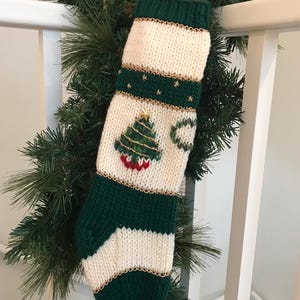 以下が含まれることがあります： 金色の縁取りと緑色のクリスマスツリーのデザインが施された、緑と白のニット製のクリスマスストッキング。ストッキングは白い手すりにかかっています。