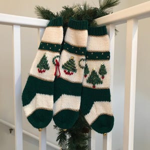 以下が含まれることがあります： 緑と白のストライプのクリスマスストッキングが3つ。刺繍されたクリスマスツリーと金色のトリムが施されています。